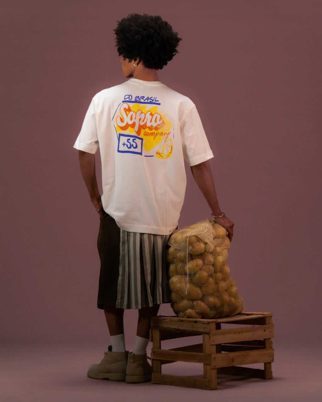 Homem de costas vestindo camiseta branca oversized com estampa colorida nas costas, segurando saco de batatas em caixa de madeira