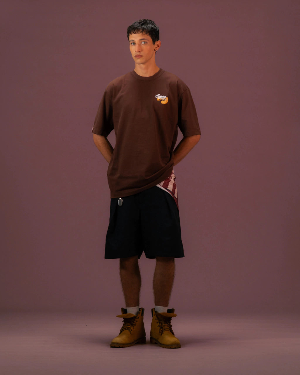 Homem vestindo camiseta oversized marrom Cajuína com estampa pequena no peito, bermuda preta e botas amarelas
