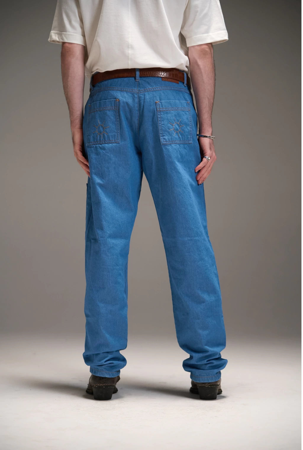 Vista traseira de calça jeans azul Aporo Sulco com bolsos quadrados bordados e cinto marrom, usada por modelo com camiseta branca e botas pretas