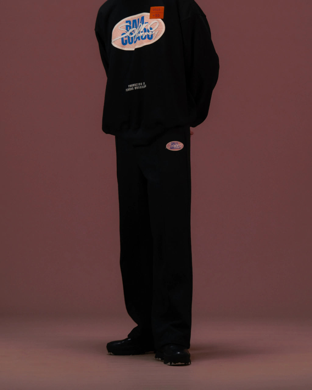 Modelo vestindo calça preta Precio com camiseta preta estampada, em fundo marrom