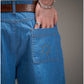 Detalhe da calça jeans azul Calça Aporo Sulco com bolso traseiro bordado com desenho de sol, usada com cinto marrom e pulseira prateada