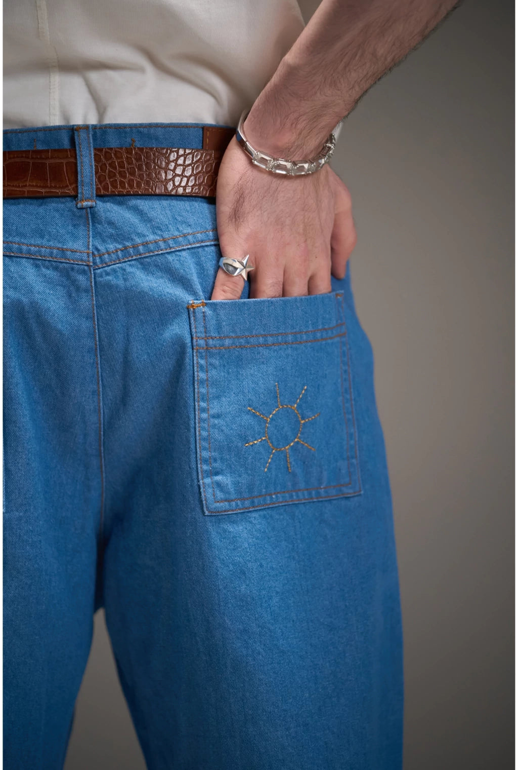 Detalhe da calça jeans azul Calça Aporo Sulco com bolso traseiro bordado com desenho de sol, usada com cinto marrom e pulseira prateada