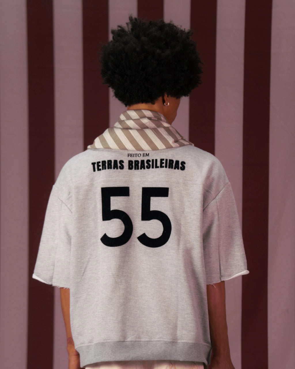 Camiseta cinza oversized com número 55 e texto "Feito em Terras Brasileiras" nas costas, usada por pessoa vista de costas.