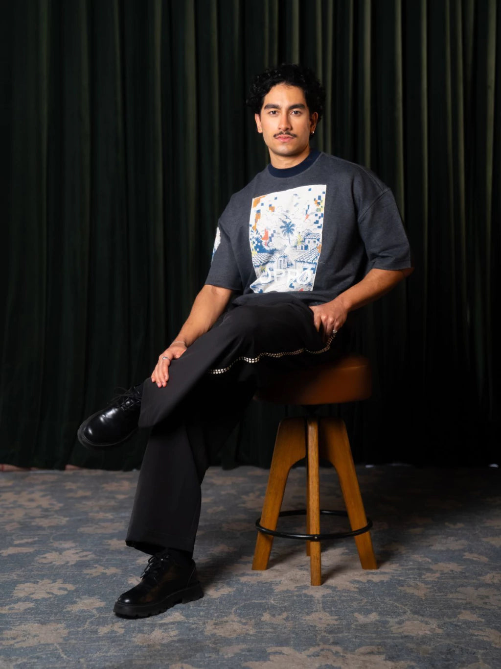 Homem sentado em banqueta de madeira usando camiseta boxy cinza com estampa de casinhas e calça preta