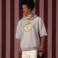 Homem usando camiseta cinza Crew Nuestra Tierra com estampa redonda e cachecol listrado, segurando bolsa preta com adesivos coloridos, em fundo de listras verticais vermelhas e rosas