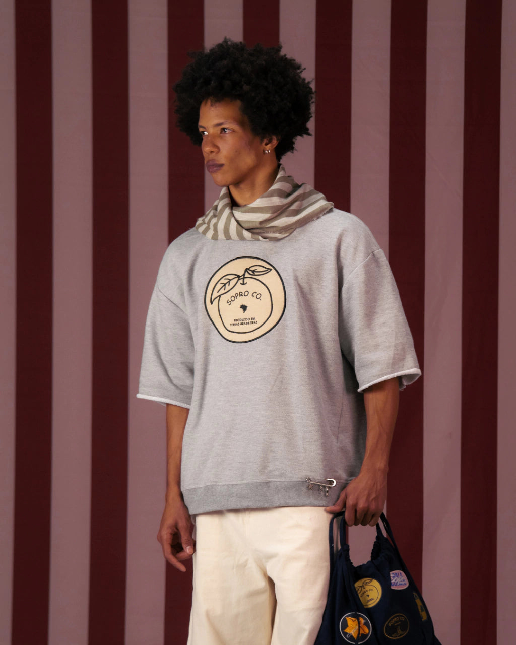 Homem usando camiseta cinza Crew Nuestra Tierra com estampa redonda e cachecol listrado, segurando bolsa preta com adesivos coloridos, em fundo de listras verticais vermelhas e rosas